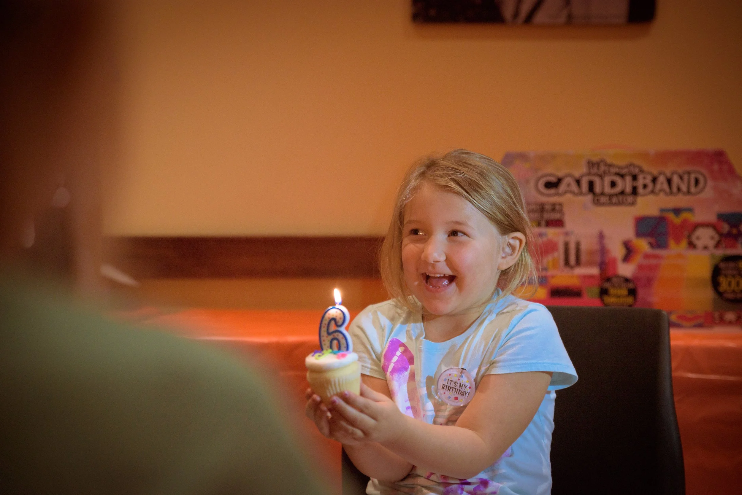 A young girl with blonde hair celebrating her 6th birthday, holding a cupcake with a lit number 6 candle, smiling. In the background, a birthday banner and a toy box labeled 'Mega Cardiband' are visible.