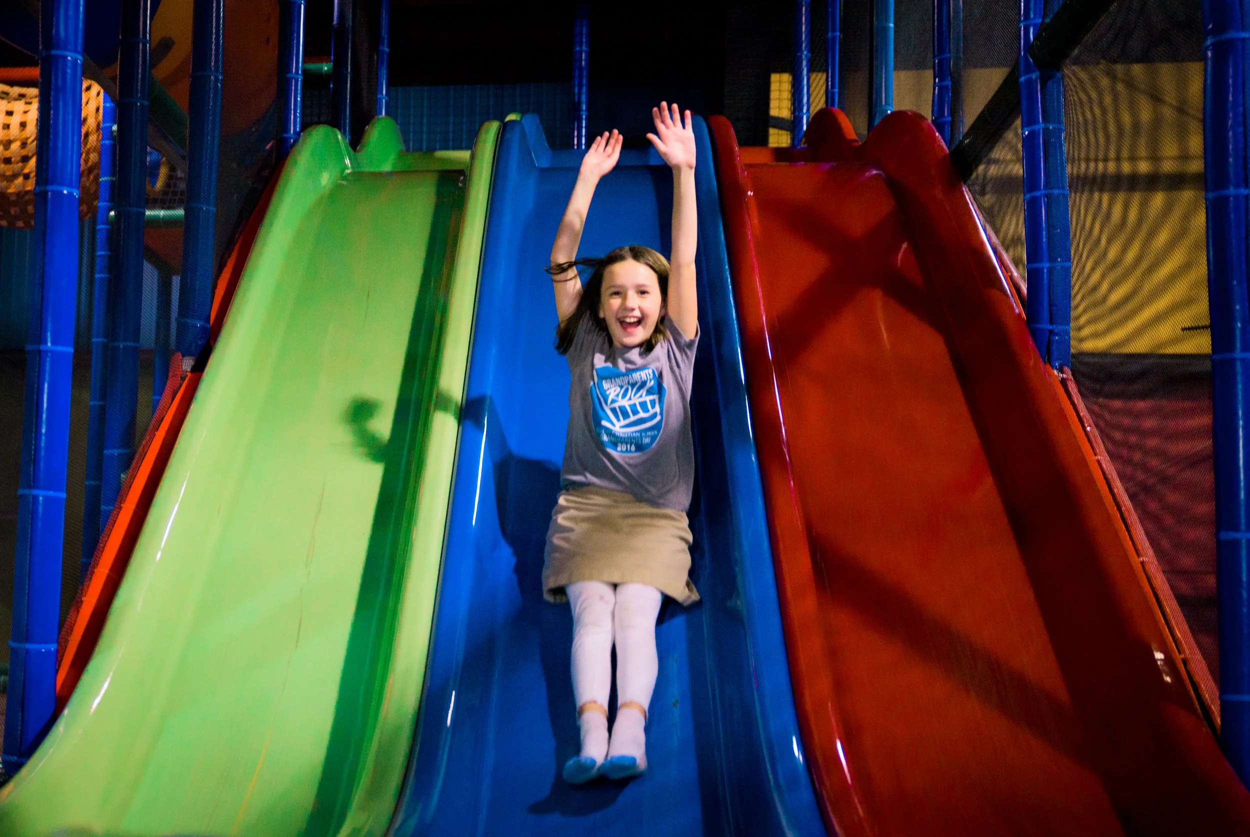 A young girl happily sliding down a colorful indoor playground slide with her arms raised in excitement.