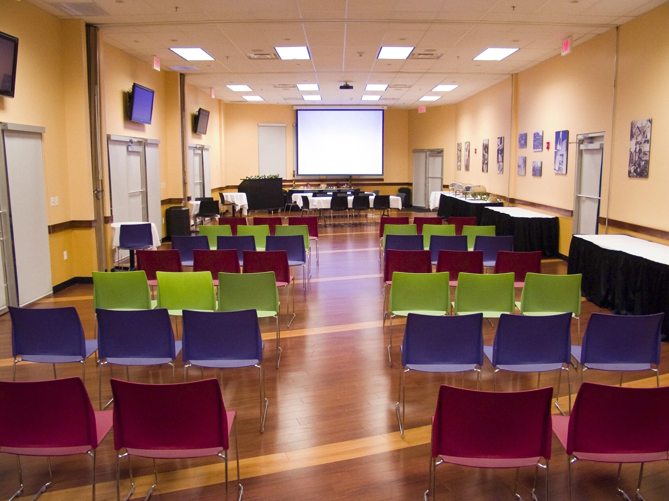 Conference room with colorful chairs arranged facing a large projection screen, with artwork on the walls and tables along the sides.