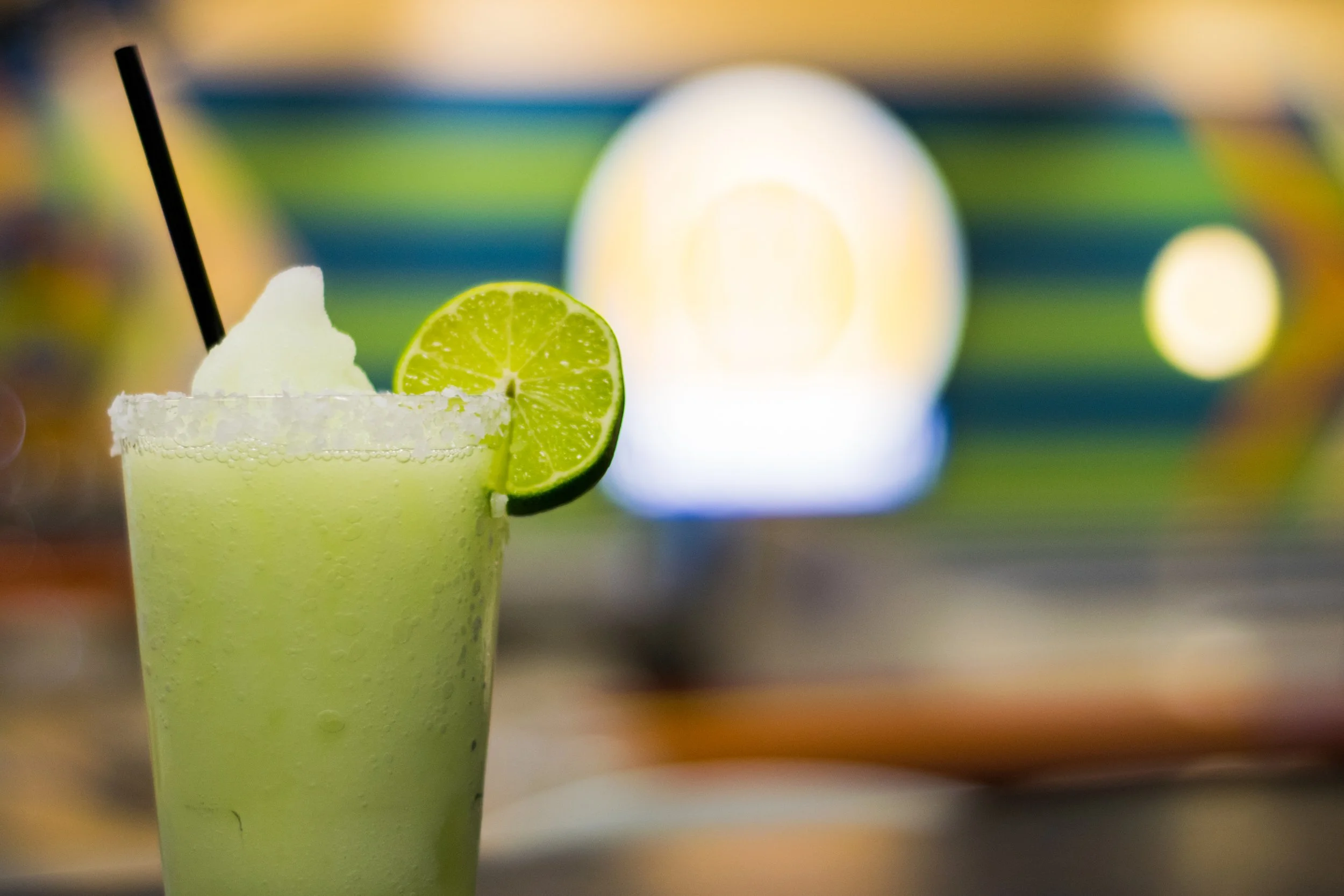 A light green margarita drink garnished with a lime wedge and a slice of lime, with a salt-rimmed glass and a black straw, out of focus colorful background.