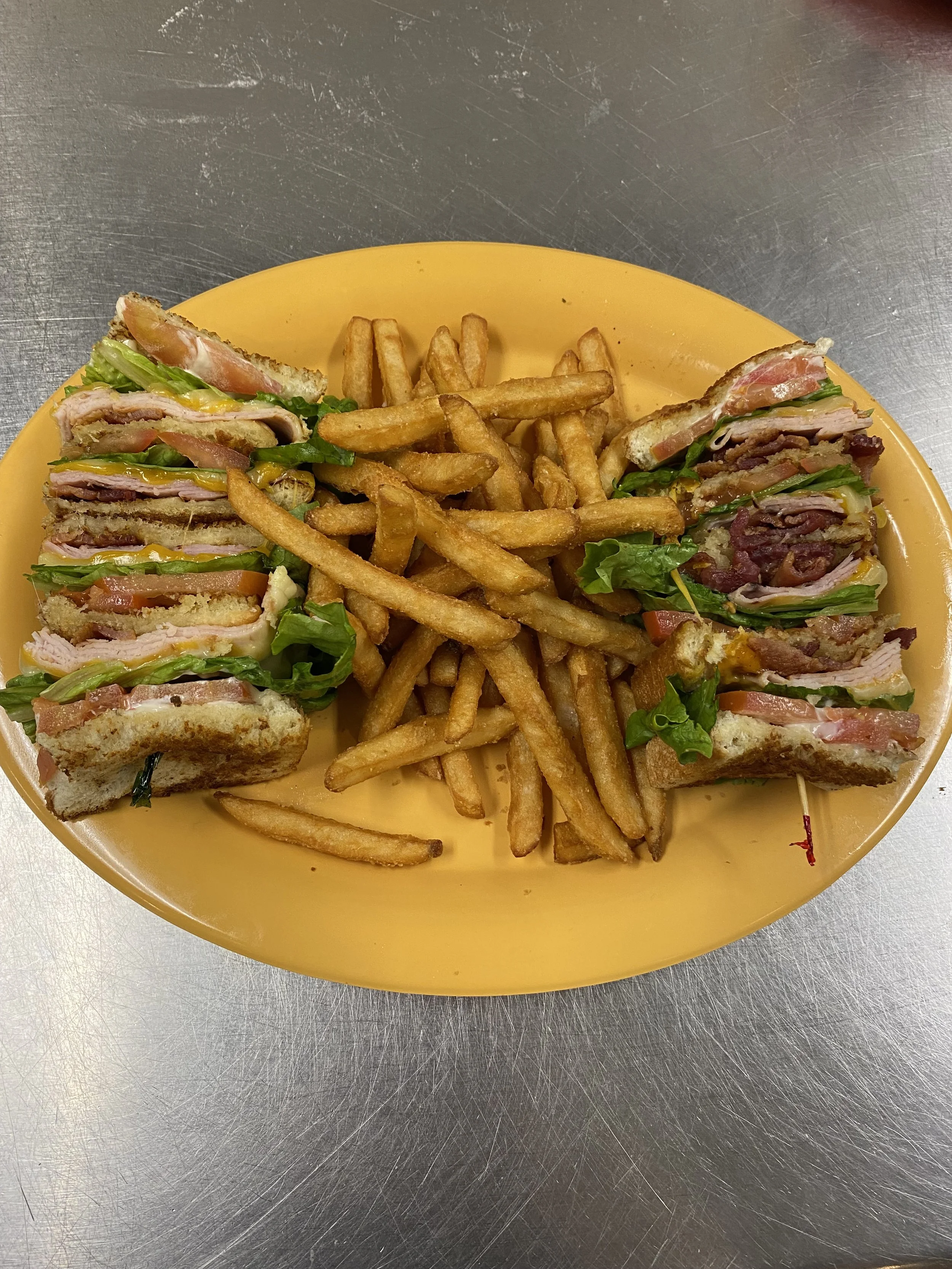 A plate with a club sandwich cut into halves on each side, filled with lettuce, tomato, bacon, and multiple layers of deli meats, with French fries in the middle.