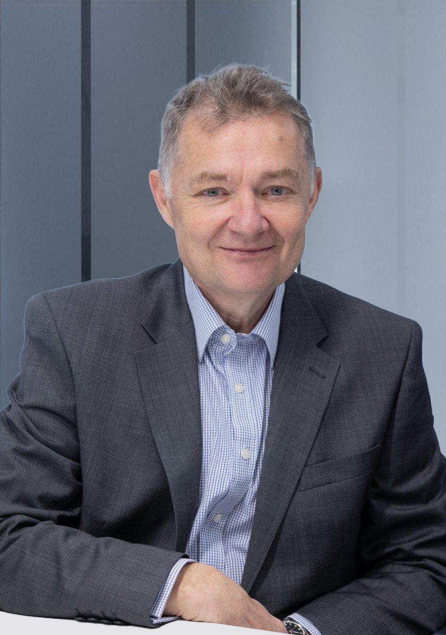 A middle-aged man wearing a dark gray suit and a light blue checkered dress shirt, smiling, sitting at a table in front of a modern, gray, paneled wall.