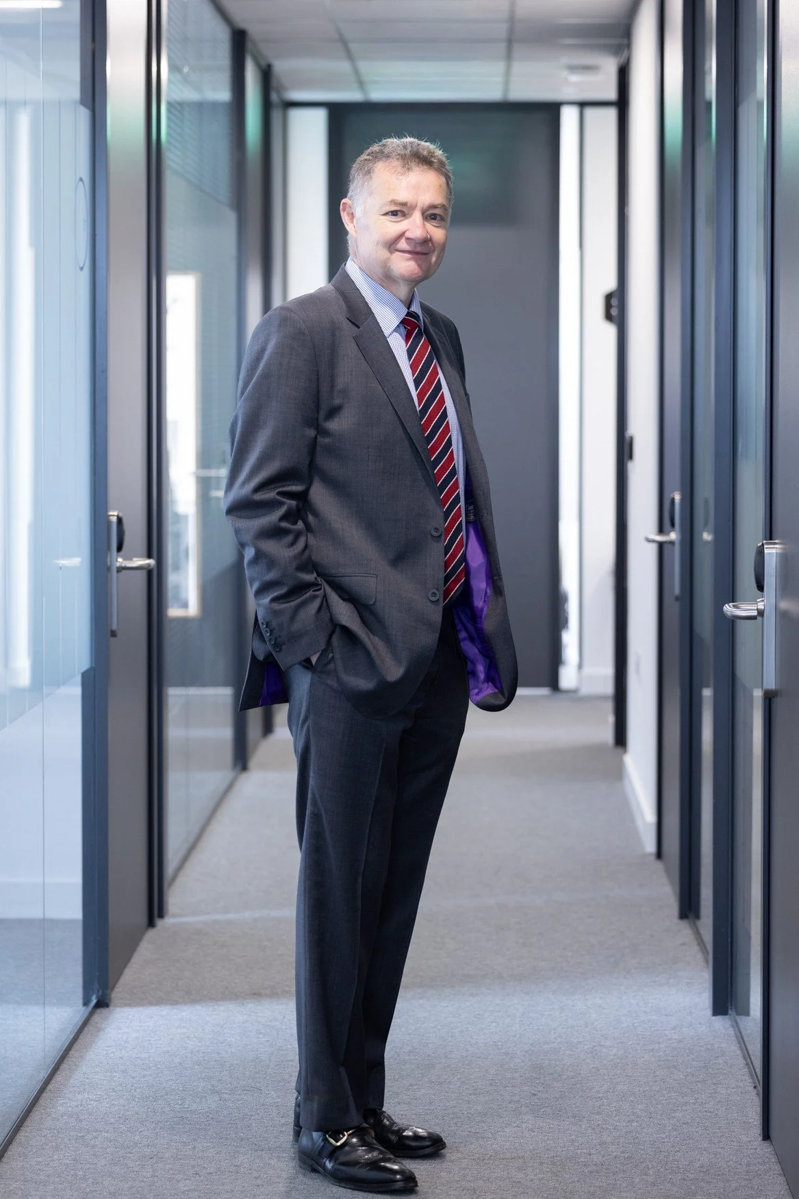 A middle-aged man in a gray suit, blue shirt, and red striped tie standing in a narrow office corridor with glass doors, hands in pockets, smiling slightly.