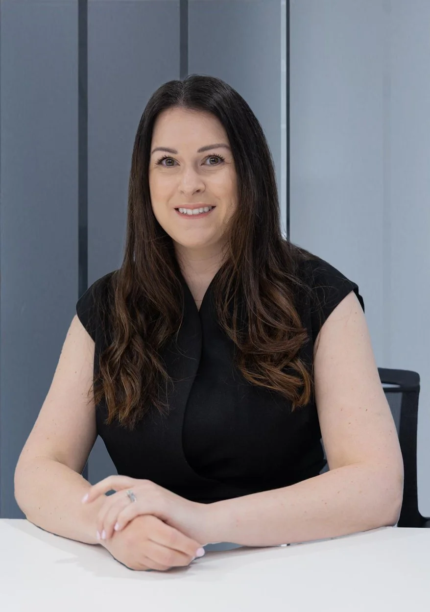 A woman with long dark brown hair, wearing a black sleeveless top, sitting at a white table, smiling, in an office setting with gray walls.