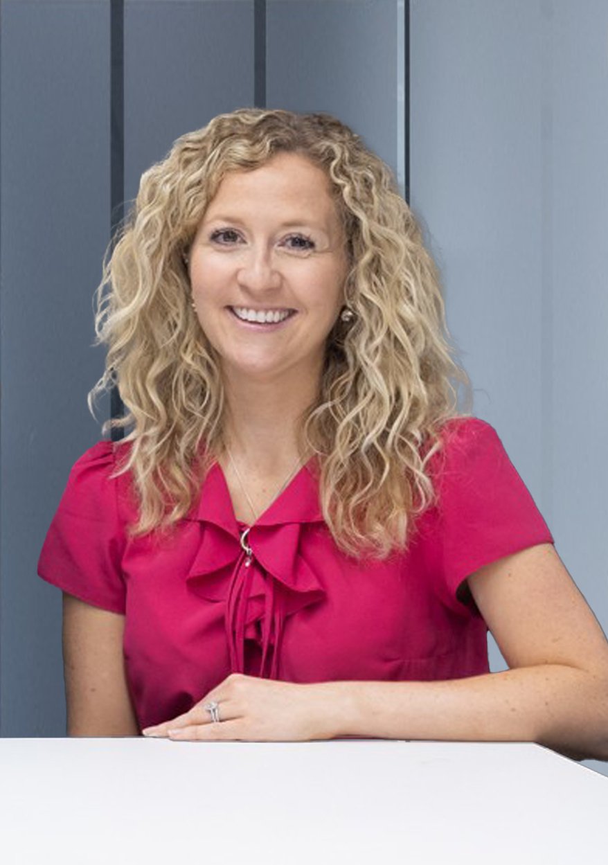 A woman with curly blonde hair wearing a pink blouse, sitting at a table, smiling, in front of a grey background.