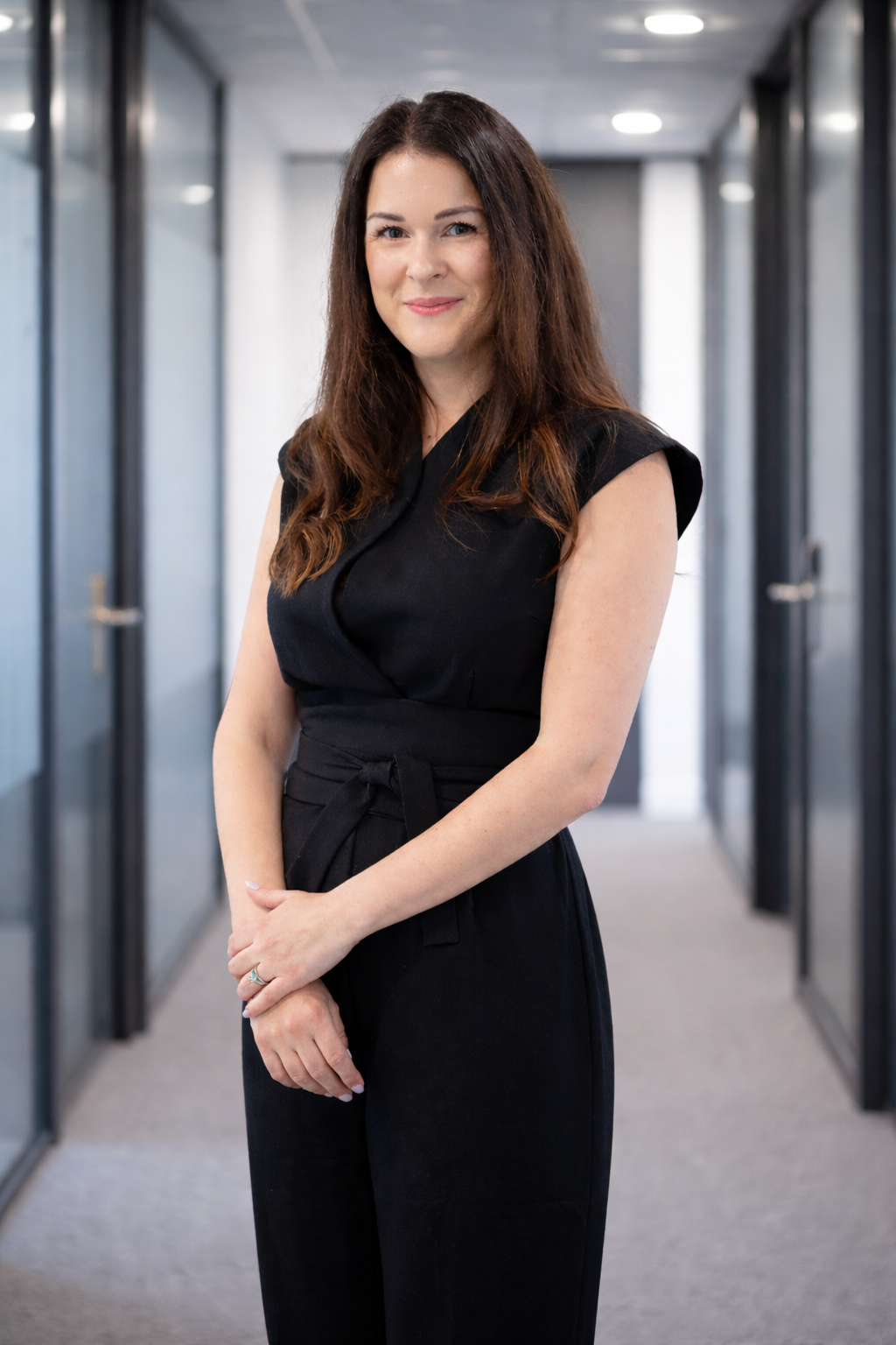 A woman with long brown hair, wearing a black sleeveless jumpsuit, standing in a modern office hallway with glass walls and doors.