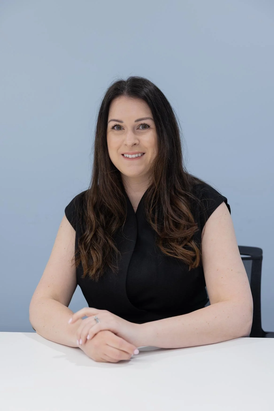 A woman with long dark hair smiling while sitting at a desk with her hands clasped. She is wearing a black sleeveless top. The background is a plain light blue wall.