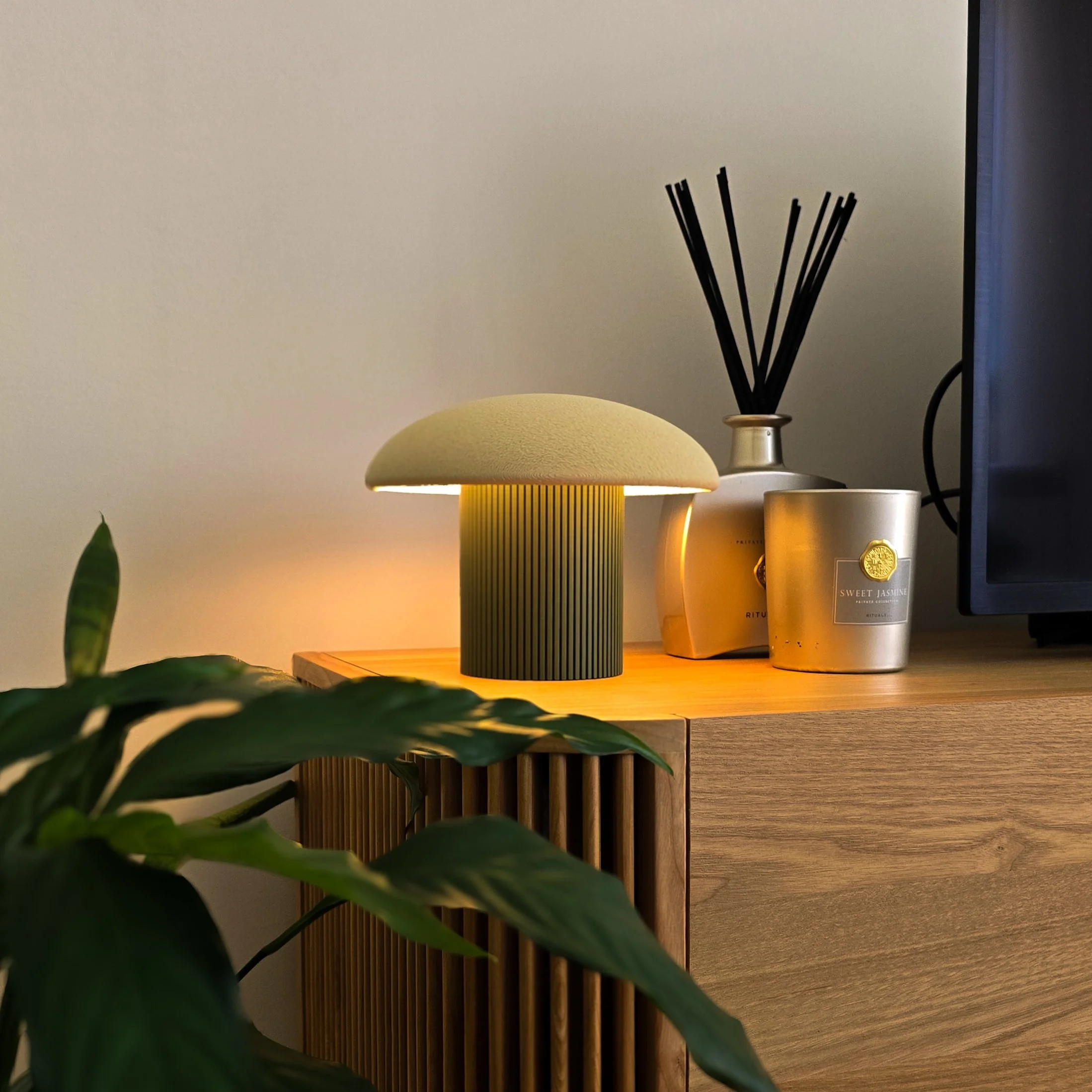 Luminária de mesa acesa com base verde e cúpula amarela em uma superfície de madeira, ao lado de vasos de cerâmica, um deles com plantas verdes, e uma parede de fundo cinza. Na borda direita, uma tela de computador preta.