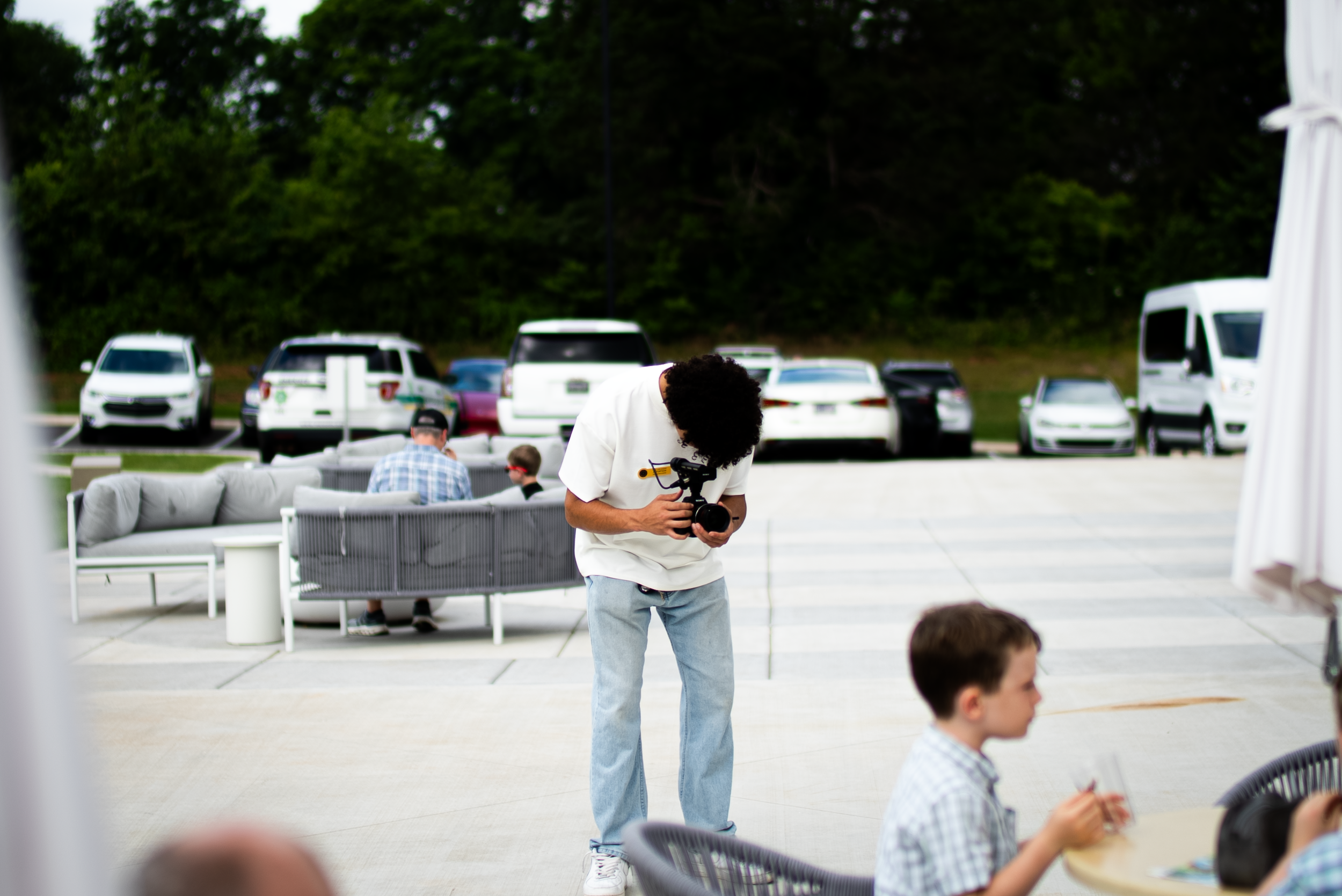 A person with curly hair wearing a white shirt holding a camera and looking down at it, standing outdoors near a table with a child sitting at it, with other people in the background and parked cars.
