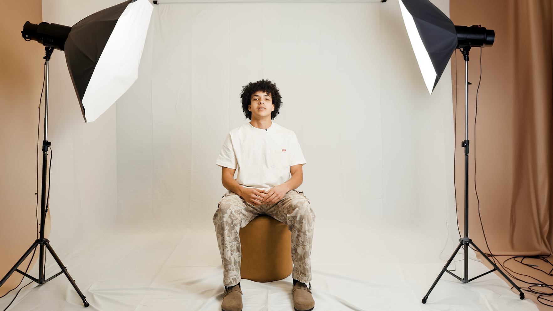 Person with curly hair sitting on a beige stool in a photography studio with a white backdrop, two softbox lights and curtains on both sides.