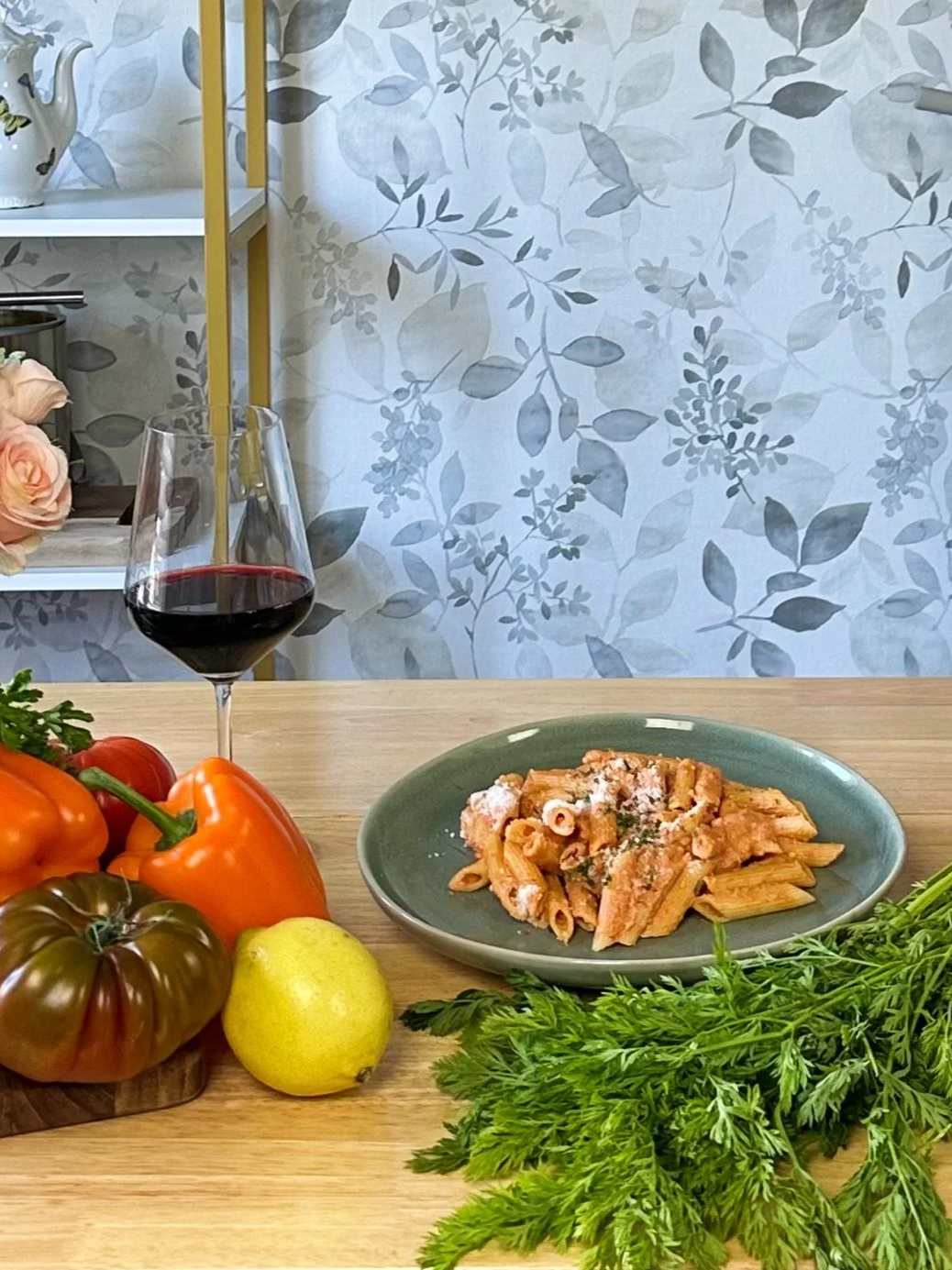 A table with a plate of pasta, a glass of red wine, colorful tomatoes, a lemon, and a bunch of leafy greens, with a background of patterned wallpaper and shelves with decorative items.