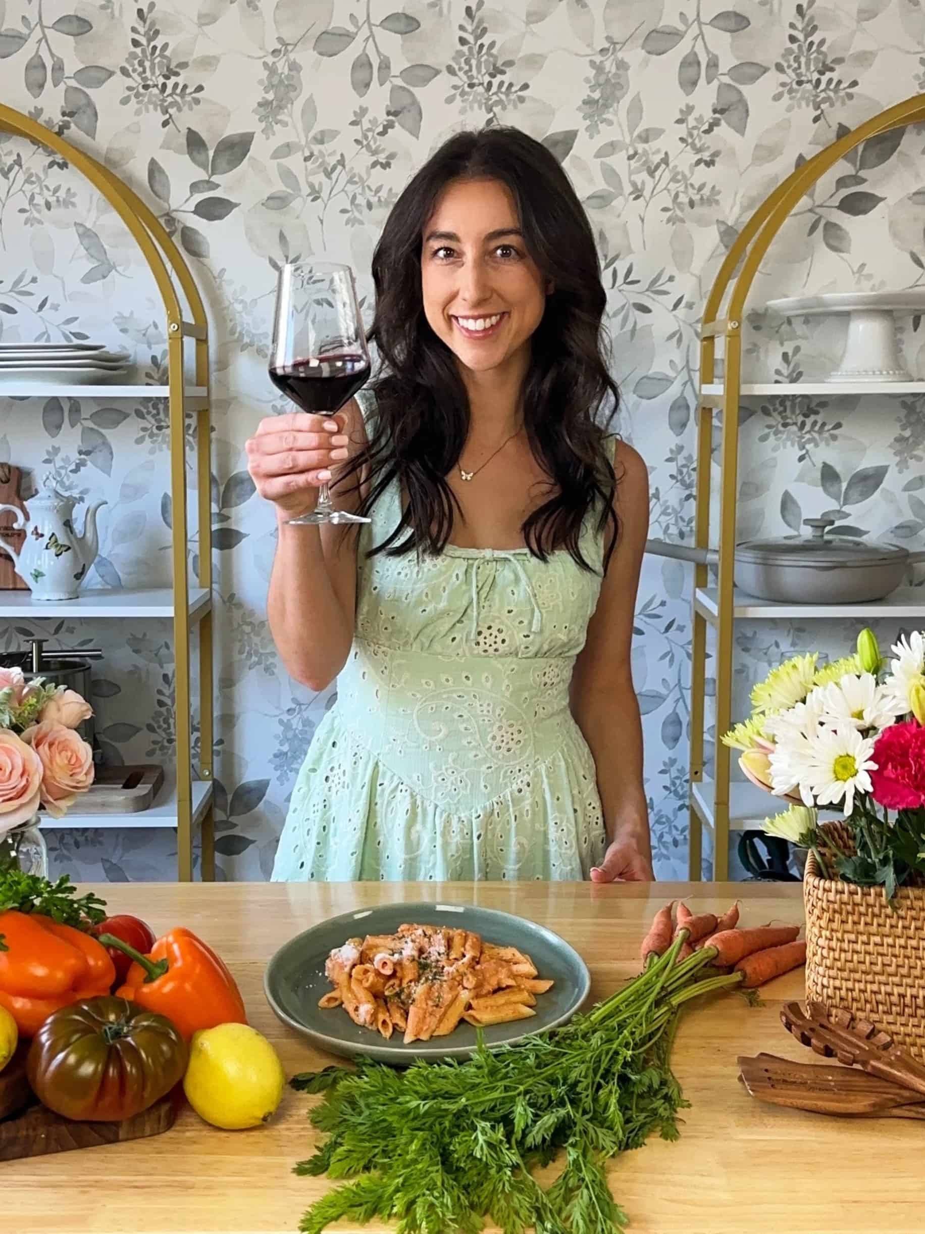 A woman with dark hair and wearing a yellow dress holds a glass of red wine and smiling at the camera. She stands behind a wooden dining table filled with colorful vegetables, a plate of pasta, and flowers in a basket. The background features patterned wallpaper and shelves with dishes and decorative items.