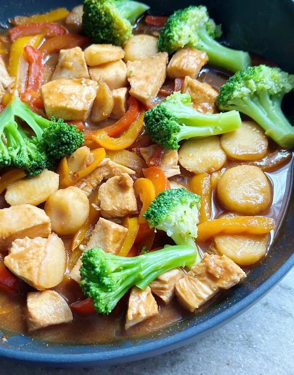 A close-up of colorful vegetable and chicken stir-fry in a black skillet, featuring broccoli, red and yellow bell peppers, sliced potatoes, and chunks of cooked chicken, all coated in a savory sauce.