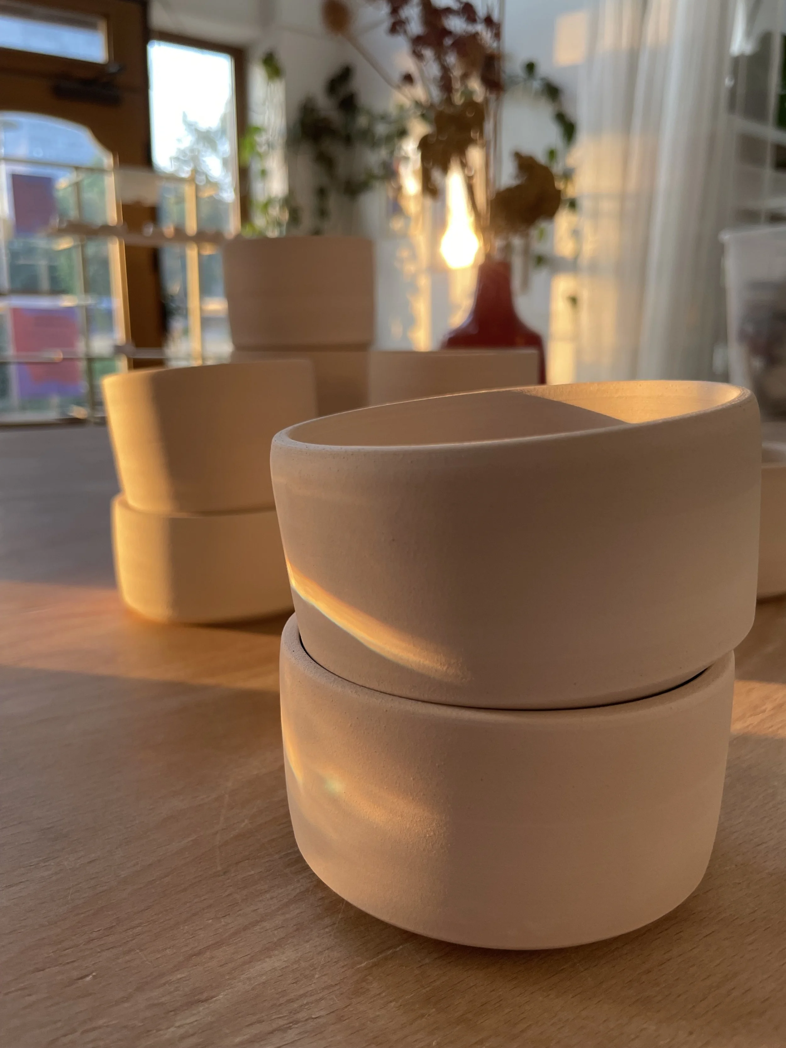 Mehrere runde Keramikbecher auf einem Holztisch, im Hintergrund eine Fensterfront mit Vorhängen und Pflanzen, der Sonnenuntergang wirft warmes Licht ins Zimmer.