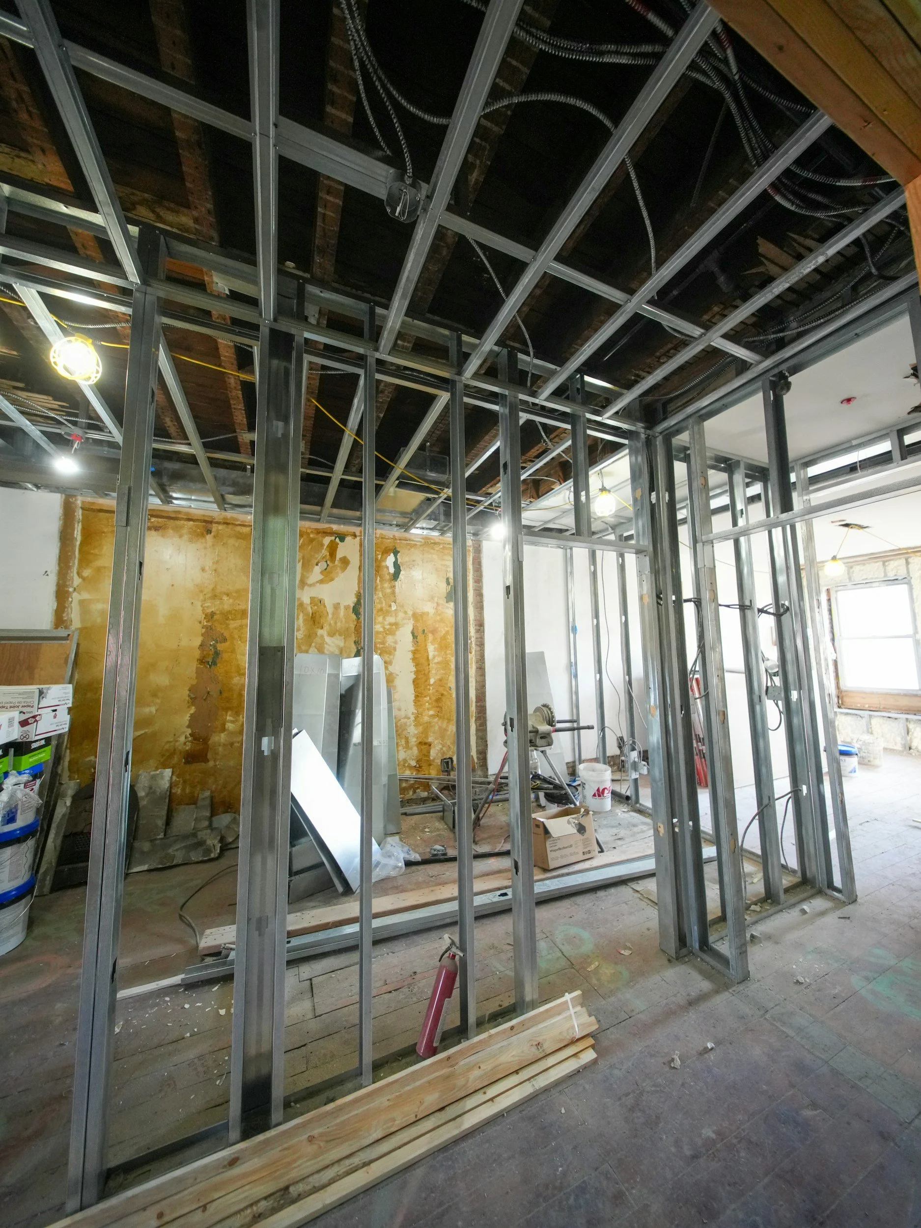 Interior view of a building under construction with metal framing, exposed ceiling, and construction tools and materials on the floor.