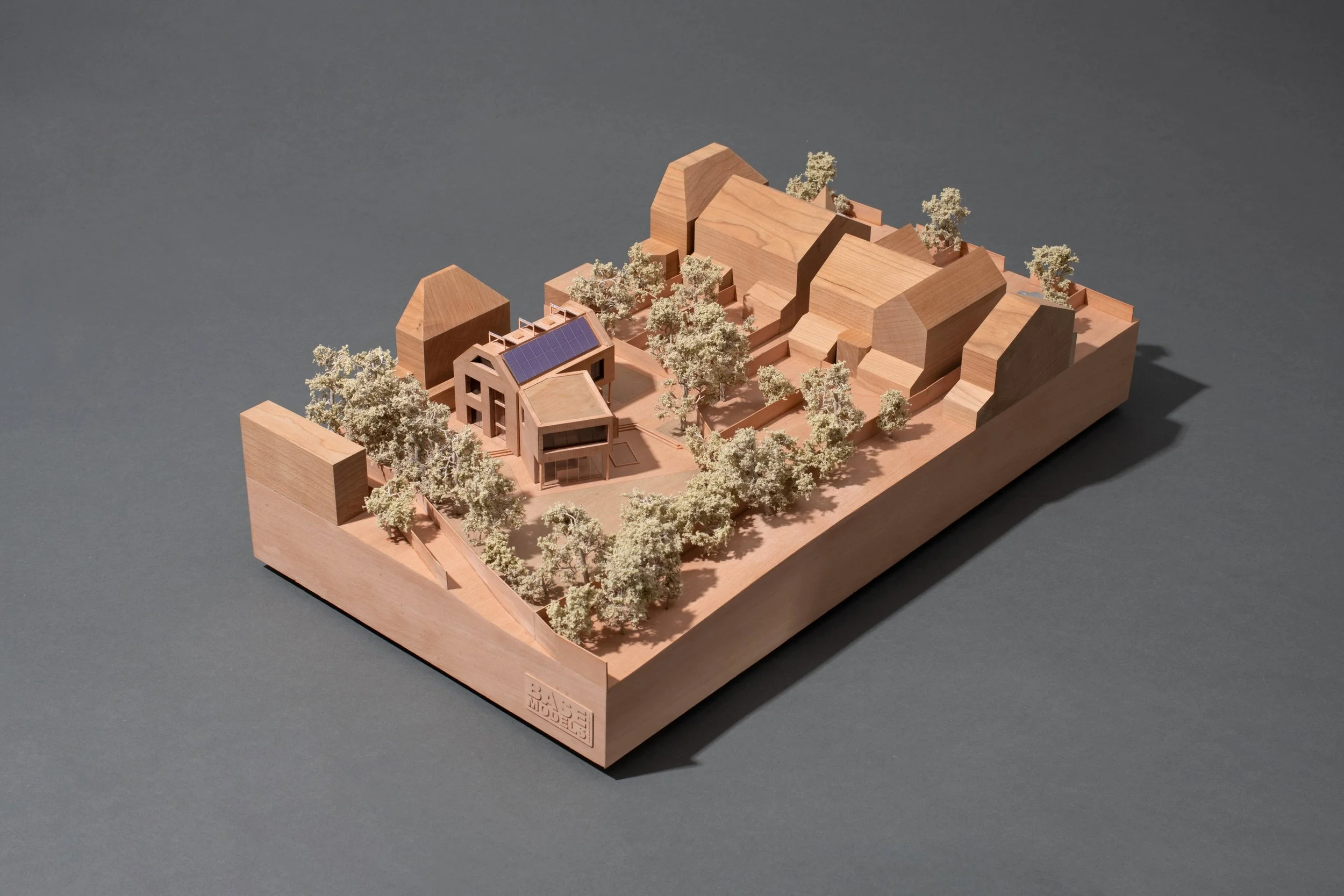 A detailed architectural model of a modern house with solar panels on the roof, surrounded by miniature trees on a wooden base.