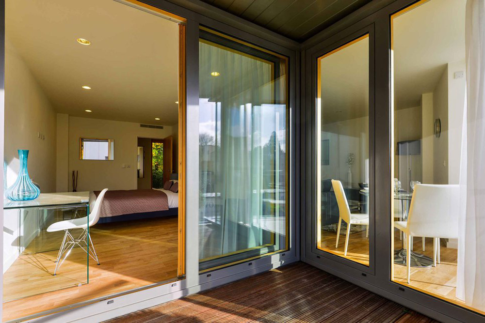 View from a private balcony looking into an NBA-designed apartment unit, showcasing hardwood flooring, minimalist furniture layout, and grey-framed structural double glazing.