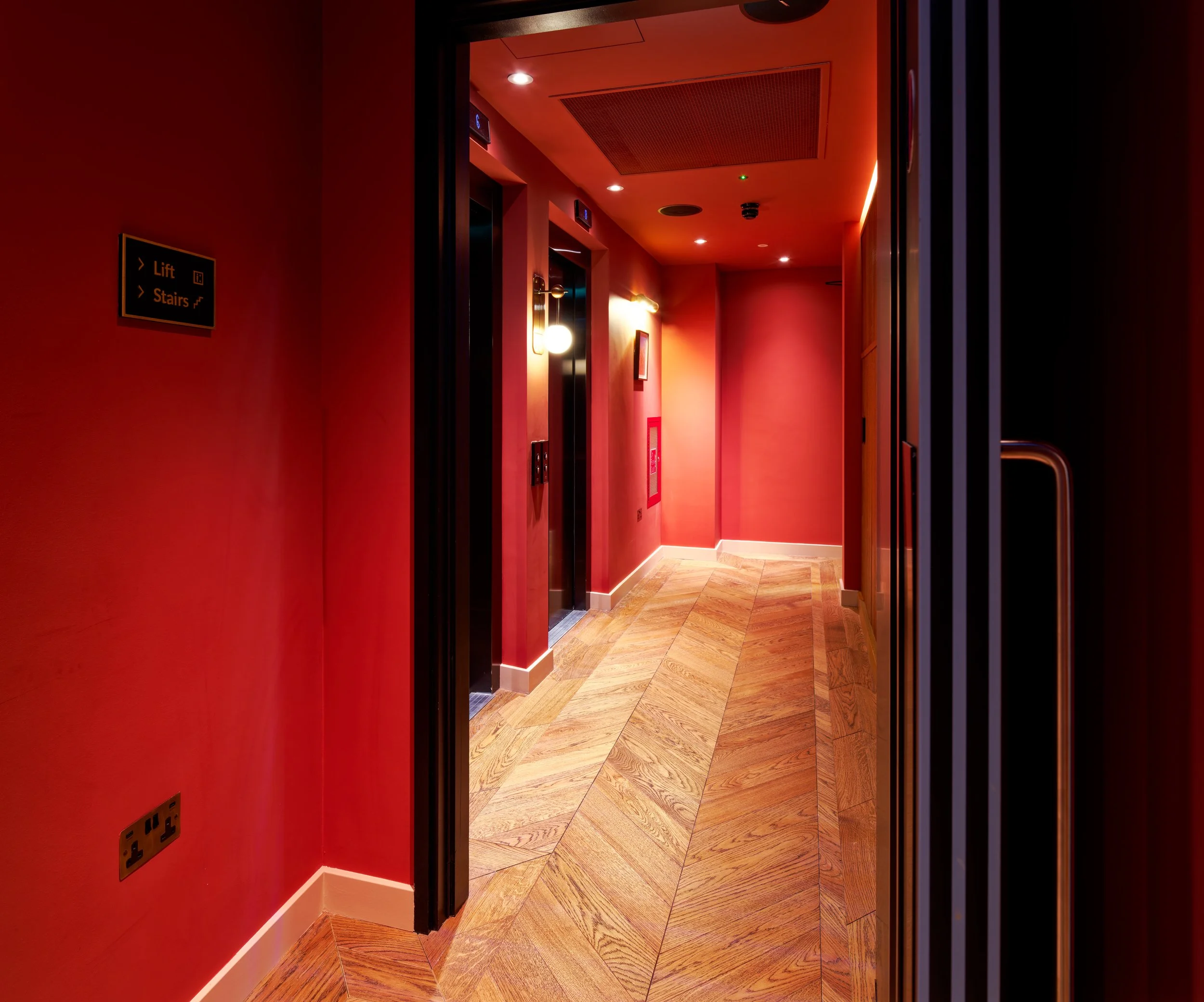 A photograph looking down a bright interior hotel corridor at the Wilde Hotel. The hallway features polished chevron wood flooring and red painted walls.
