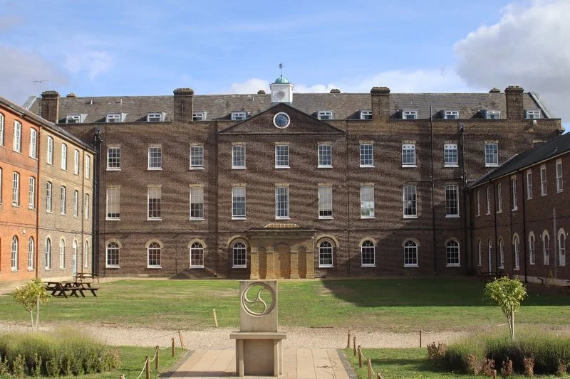 A large brick building with multiple windows, surrounded by a grassy courtyard with trees and a sculpture in the foreground.