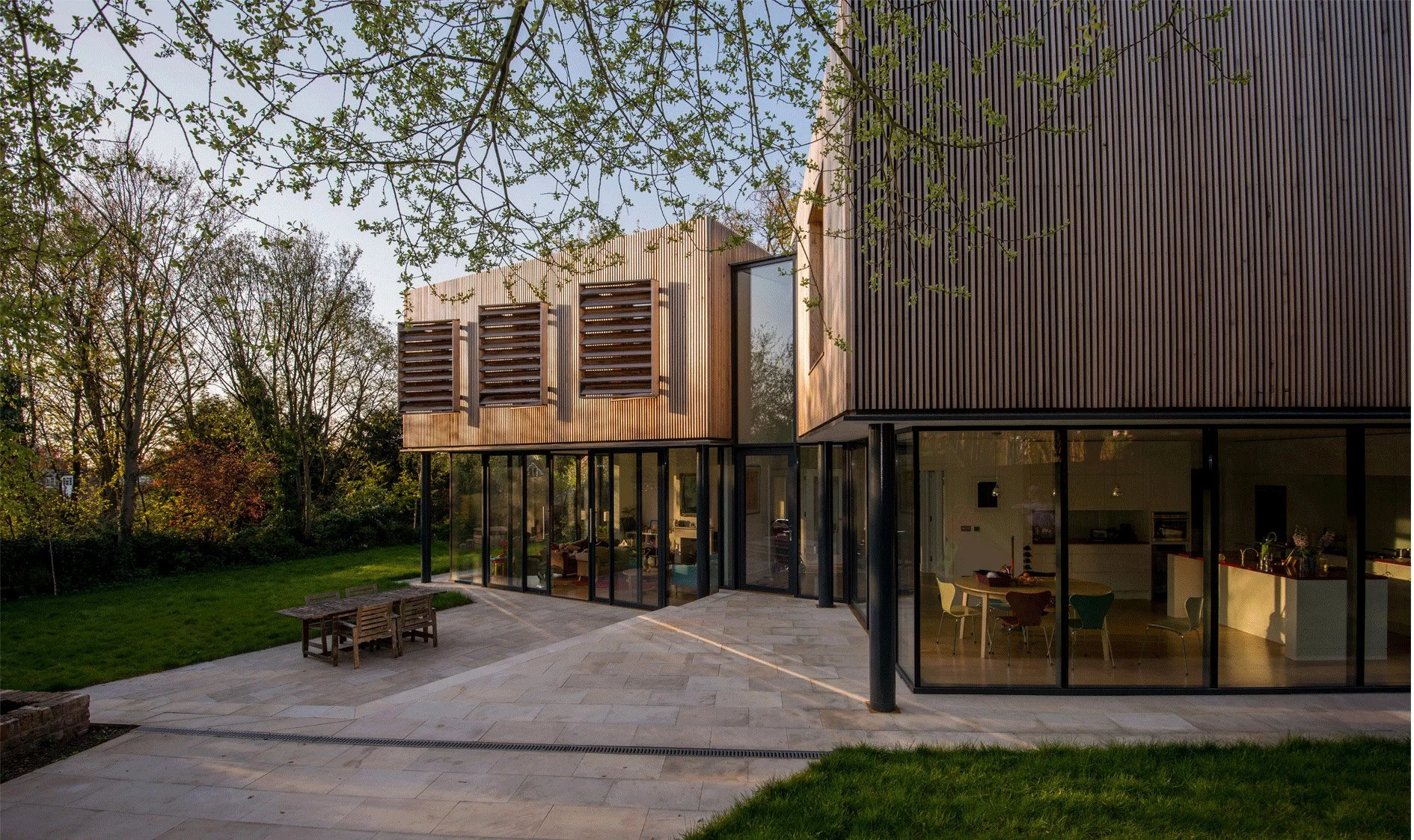Nick Baker Architects — Modern house with wooden and dark panel exterior, large glass windows, and an outdoor patio with a wooden table and chairs, green lawn, trees, and a c