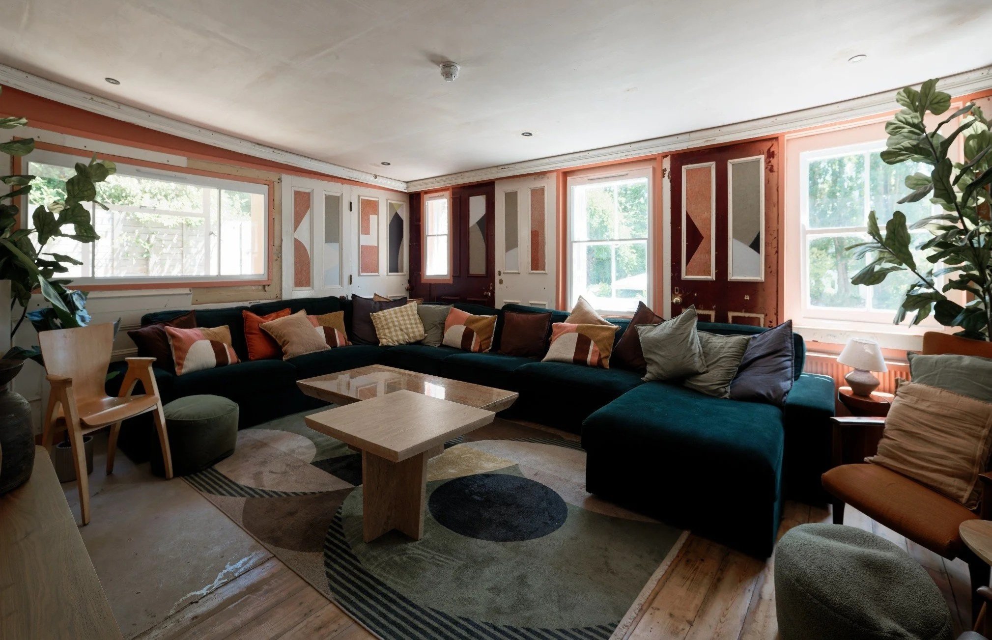 Living room with large windows, sectional sofa with multiple pillows, wooden coffee tables, plants, and a patterned rug.