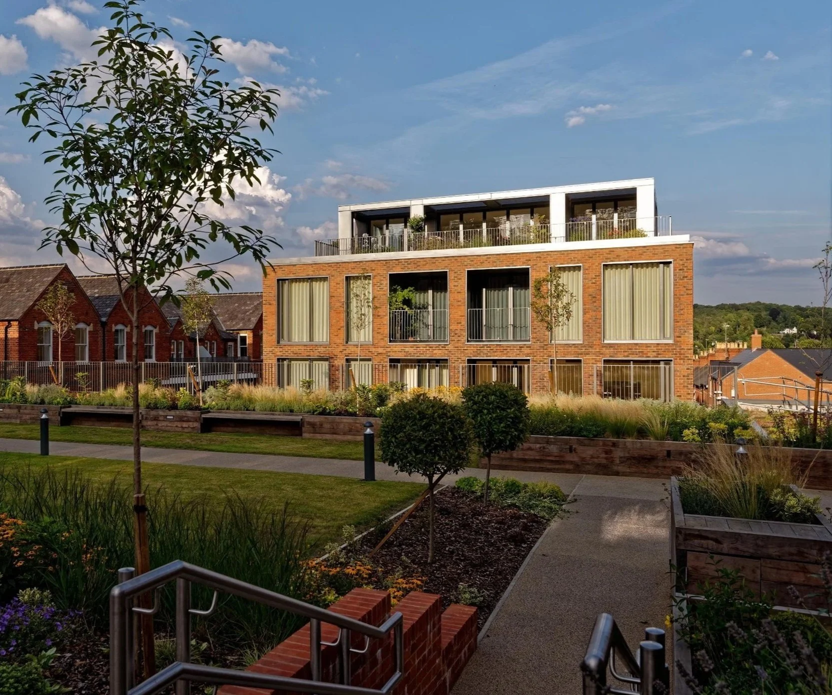 View of modern residential buildings with brick and glass facades, surrounded by landscaped gardens, trees, and walking paths under a partly cloudy sky.