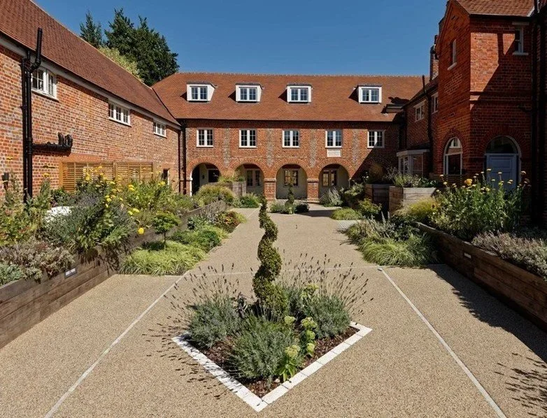 A courtyard garden surrounded by brick buildings with red-tiled roofs, featuring a central gravel pathway, flower beds with various plants and shrubs, and a tall, spiral-shaped shrub in the middle.