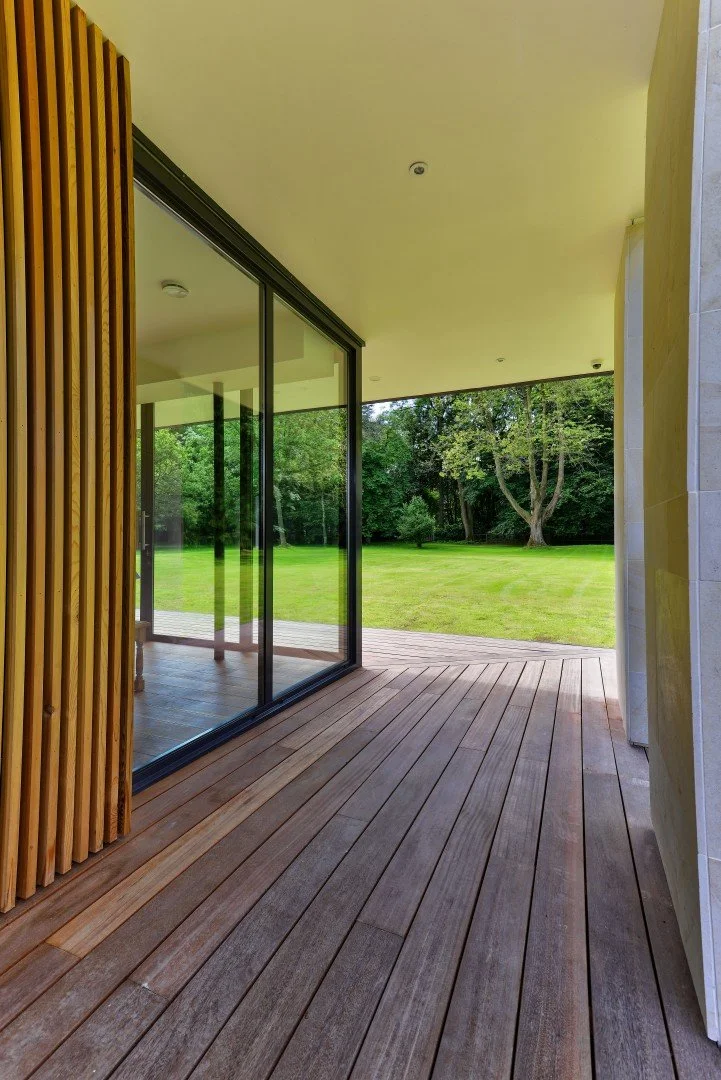 View from a covered timber deck terrace of a modern pavilion house, featuring large sliding glass doors, vertical timber fins, and a limestone pier looking out over a large green lawn and mature trees.