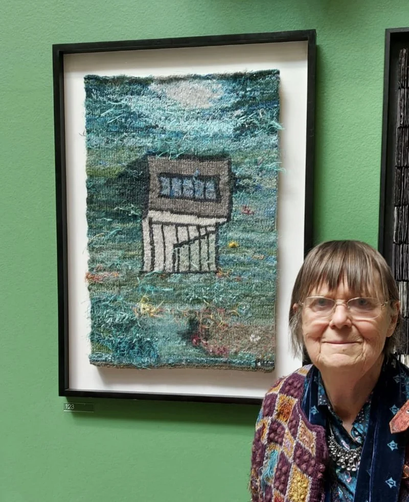 A woman with glasses and a colorful sweater stands in front of a framed textile artwork depicting a house with an upstairs window and a staircase, against a green wall background.