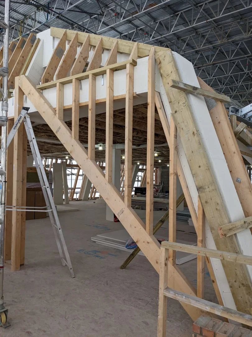 A close-up site photograph focusing on a section of the steel skeleton being lined with vertical timber stud framing and insulated infill panels.