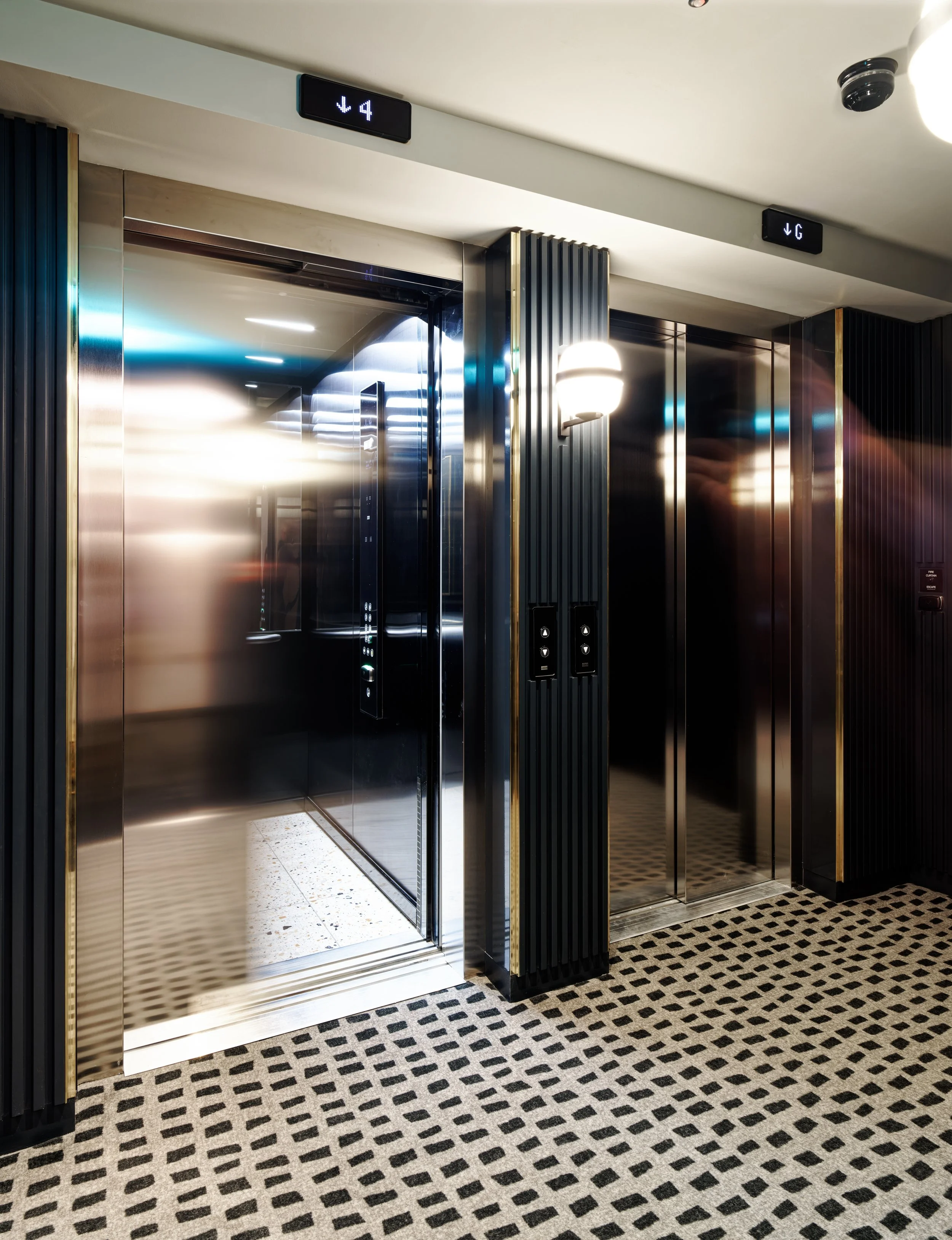 A modern hotel lift lobby featuring two polished stainless steel elevator doors framed by dark fluted wall paneling and elegant gold trim.