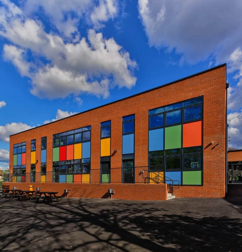 A modern brick school building with large, colorful window panels under a partly cloudy sky.