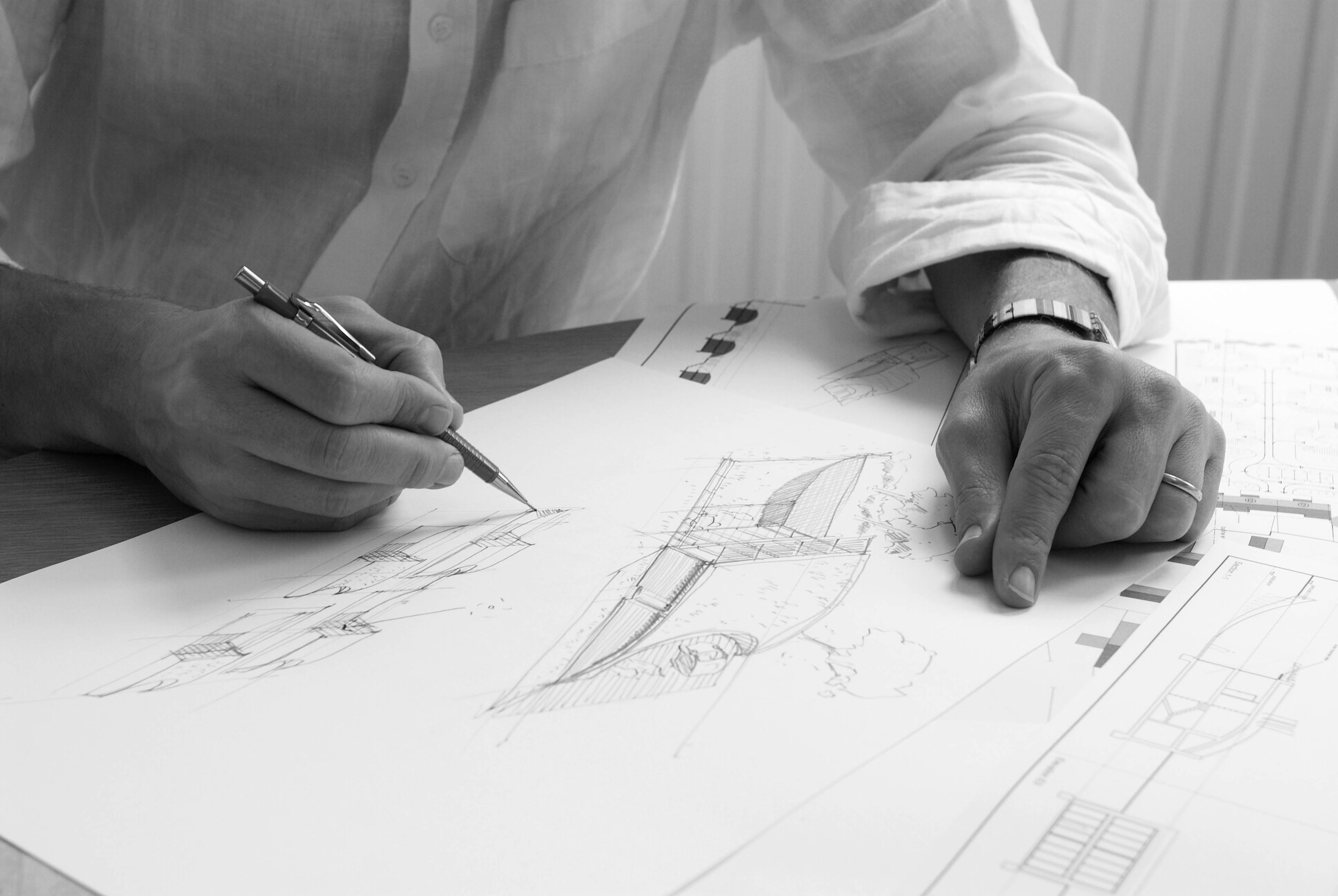 A person wearing a white long sleeve shirt and a wristwatch is drawing blueprints or architectural sketches on paper with a pen.