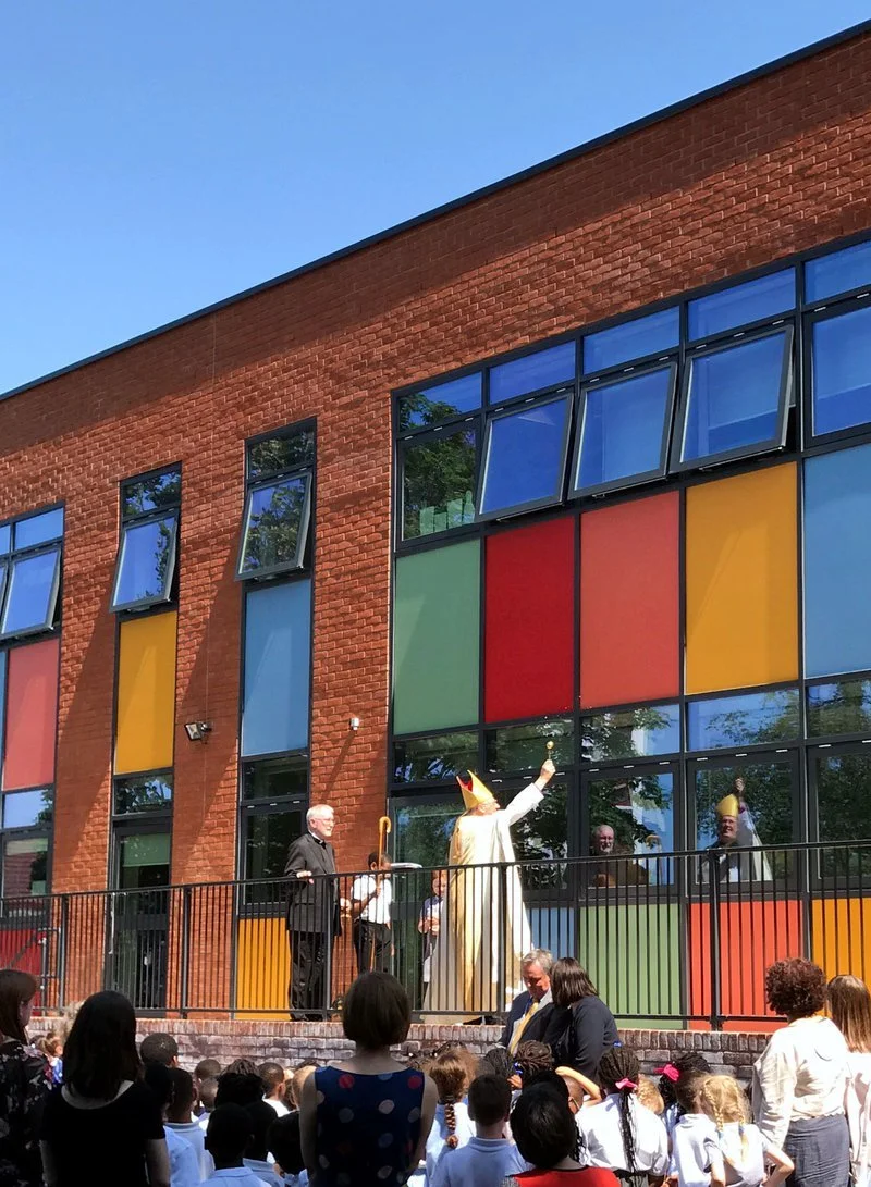 A clown and several people on an outdoor stage in front of a colorful, stained-glass style building with children watching.
