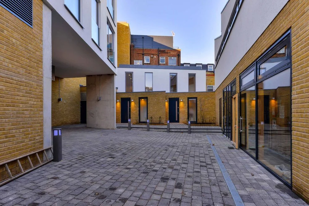 Architectural photograph of the completed Delancey courtyard at Solstice Point designed by NBA, showing a paved open courtyard surrounded by yellow brick facades, glass storefront entries, and structural concrete pillars.