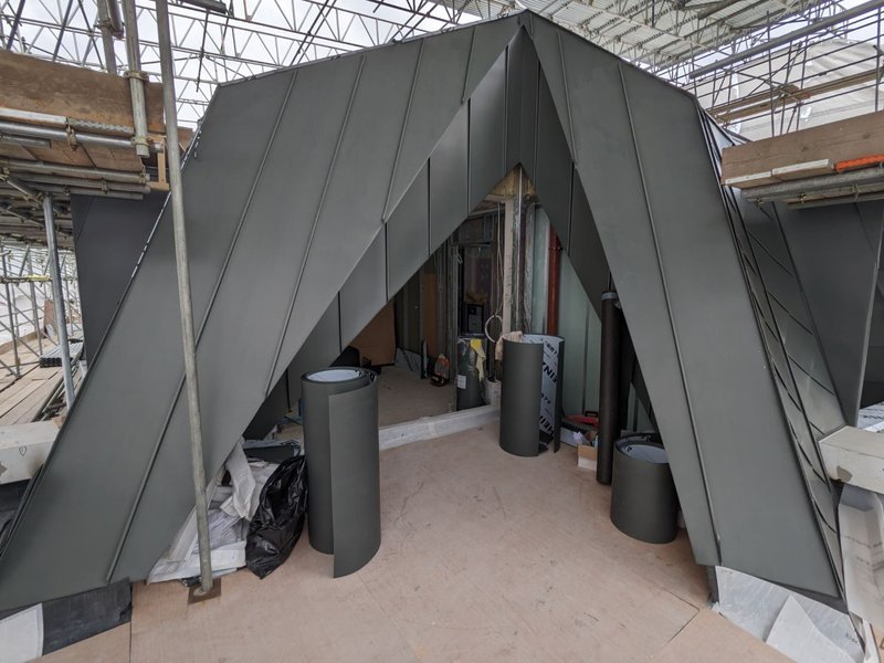 Construction site with a modern, angular black metal structure and three cylindrical black pillars inside. The area is under construction with scaffolding and building materials visible.