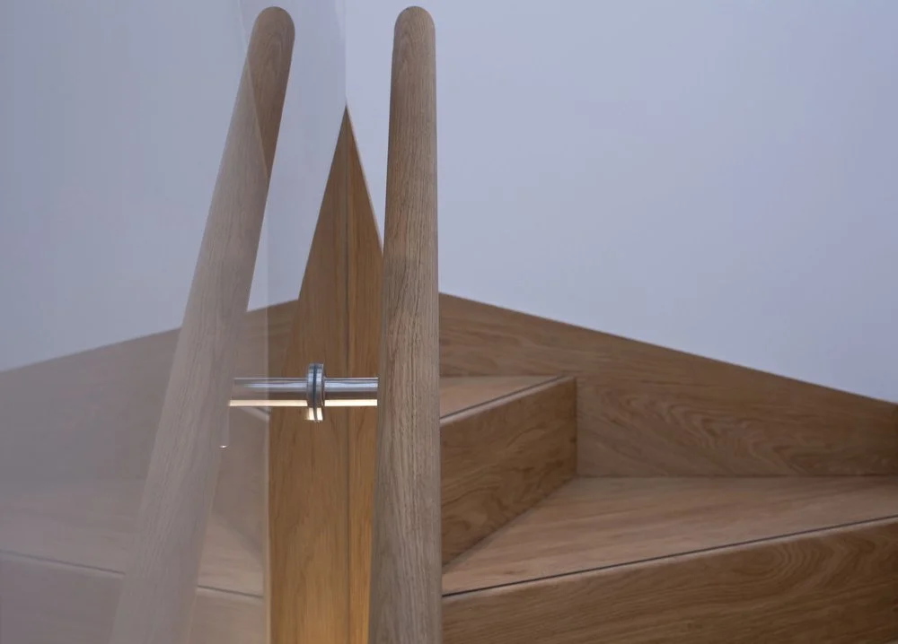 Close-up architectural shot of a contemporary wooden staircase, highlighting the sharp clean lines of oak winders meeting vertical timber columns behind a clear glass safety panel secured by metallic hardware.
