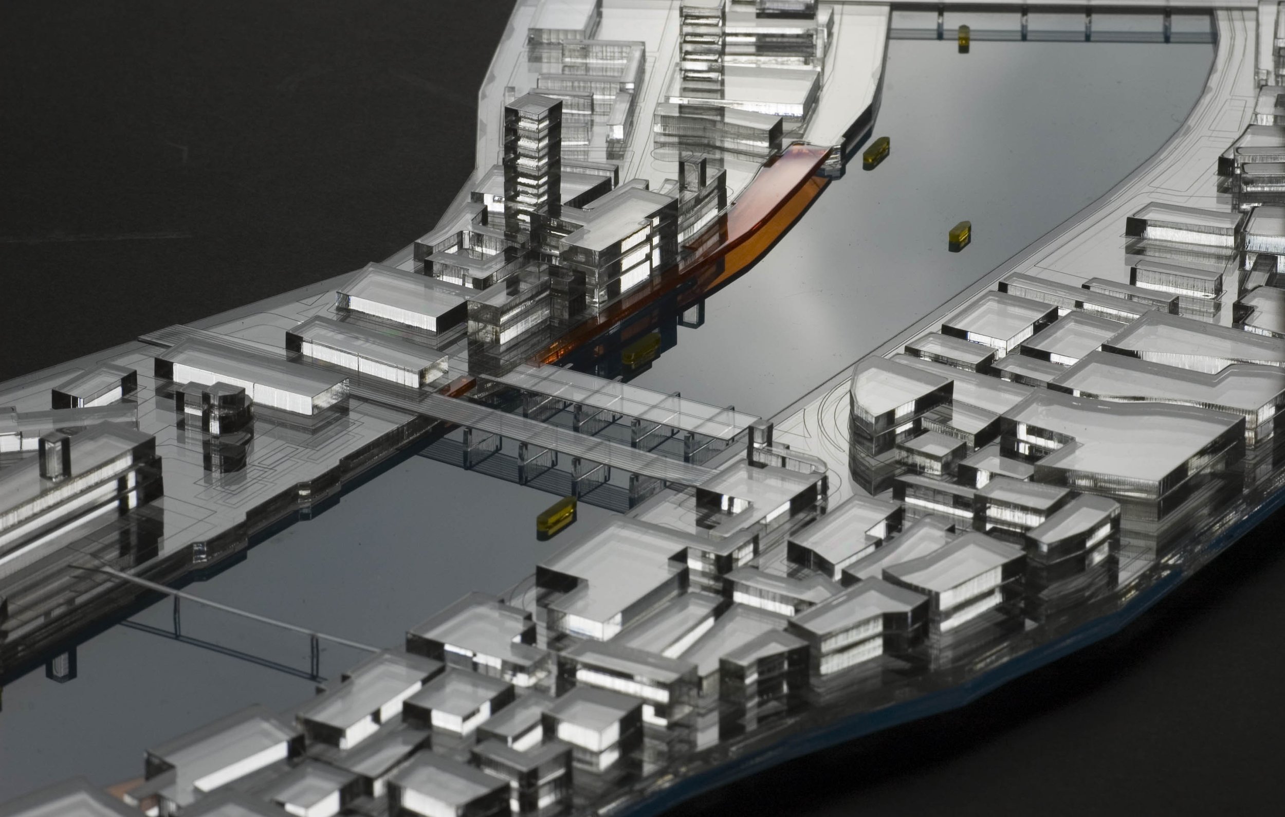 A detailed architectural scale model of a modern urban development, featuring high-rise buildings, roads, and water body, made from transparent and reflective materials.