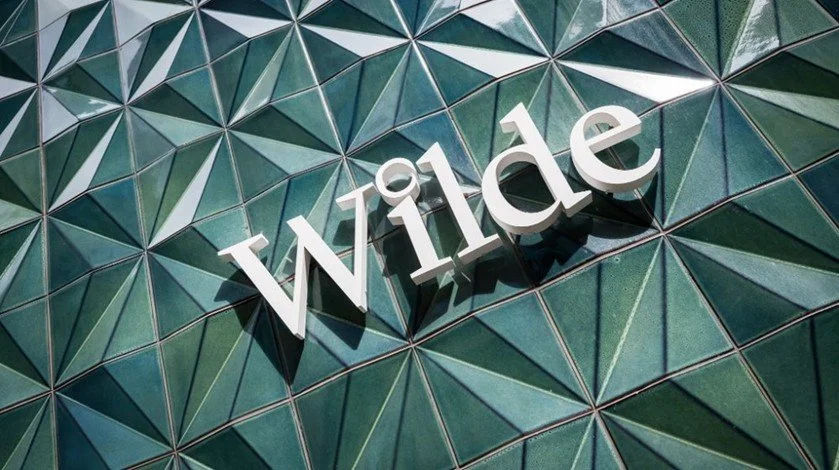 The word 'Wilde' in white 3D letters mounted on a geometric glass building facade with green-tinted panels.