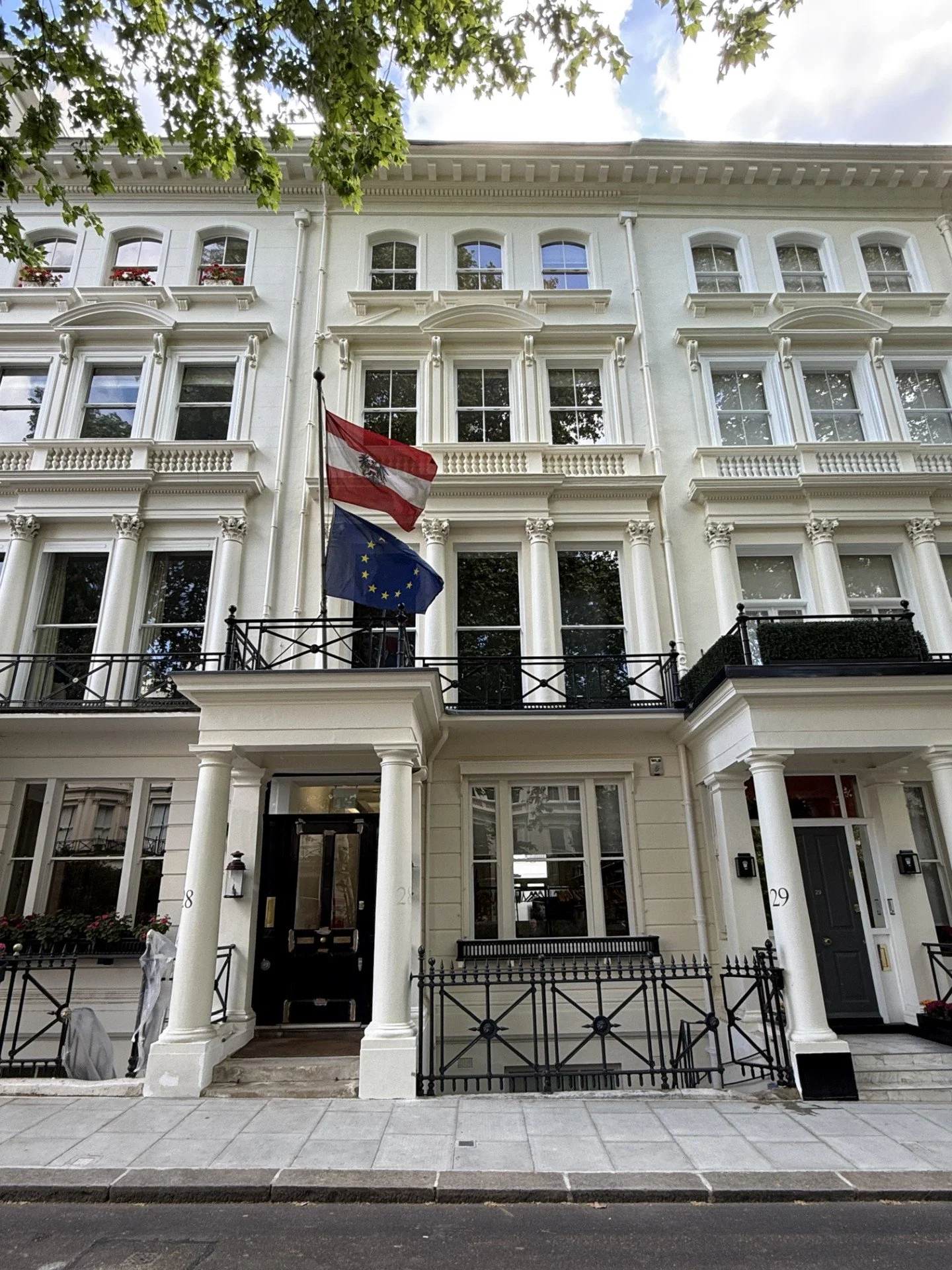 White multi-story building with ornate architectural details, black railings, and flags of Austria and the European Union on the balcony, with trees and a blue sky with clouds above.