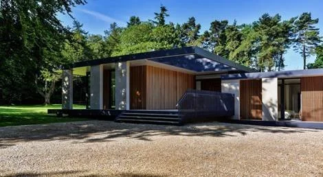 Modern house with wooden and concrete exterior, surrounded by greenery and trees, under a clear blue sky.