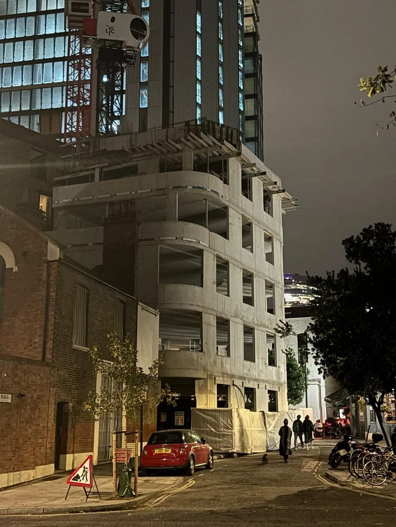 Nighttime scene of a construction site with a tall building under construction, scaffolding, and a camera or measuring device mounted on the building. In the foreground, there is a brick building on the left, a red car parked on the street, some trees, bicycles, and pedestrians walking nearby.