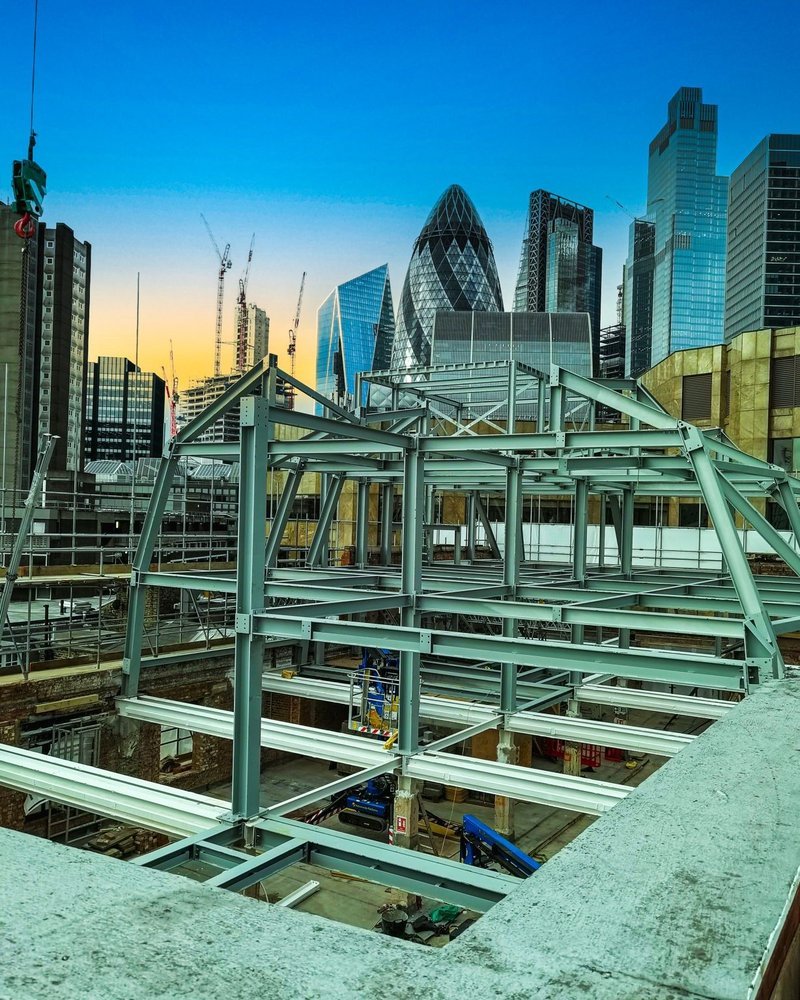 Construction site with steel framework structure in foreground and modern skyscrapers in the background during sunset in an urban city.