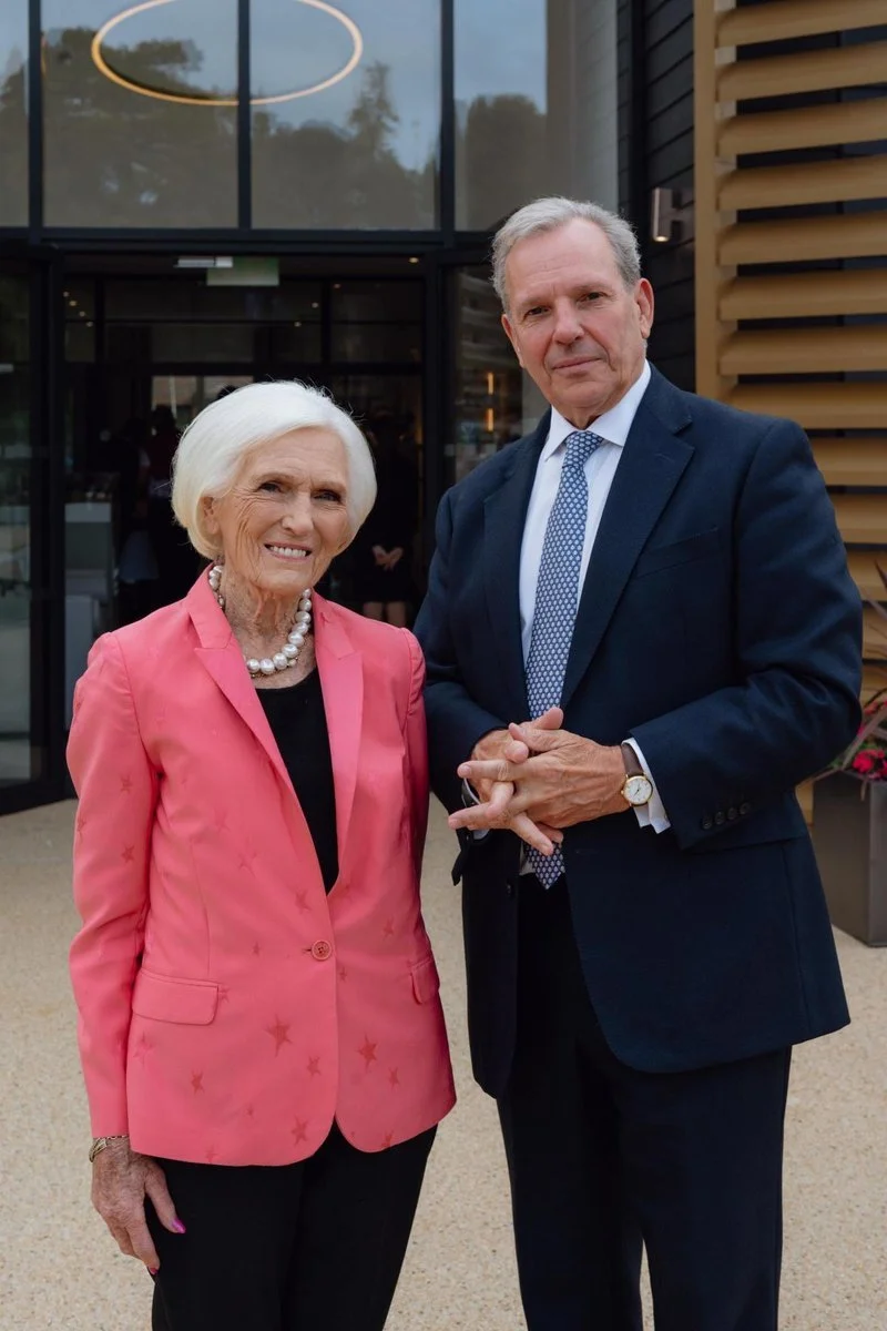 An elderly woman with white hair wearing a pink blazer with star patterns, pearls, and black pants standing next to a middle-aged man in a dark suit, light blue dress shirt, and patterned tie, outside a modern building with large glass windows and wood accents.