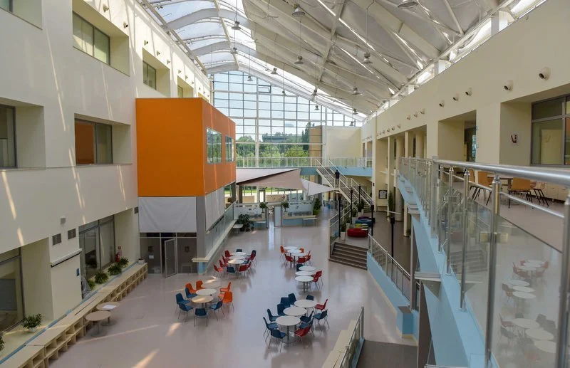 Modern indoor atrium with seating area, glass ceiling, and large windows, featuring white, orange, and blue accents.