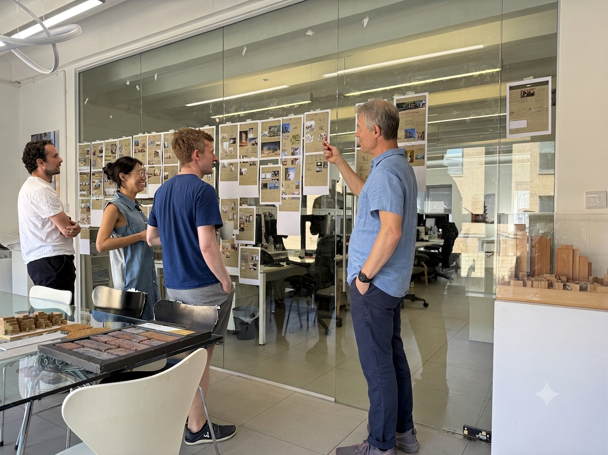 A group of four people standing inside an office, looking at a display of papers and images on a glass wall. One man is gesturing towards the display while talking. The office has chairs, a glass table with samples, and a large window in the background.