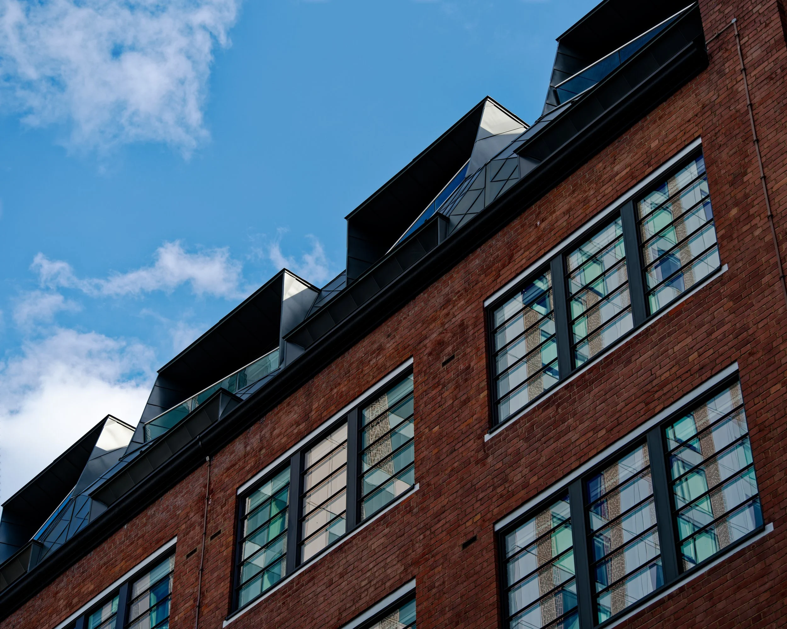 The image highlights the contrast between the traditional red brick walls with large rectangular multi-pane windows and the contemporary dark gray zinc-clad, faceted dormer windows on the roof extension.