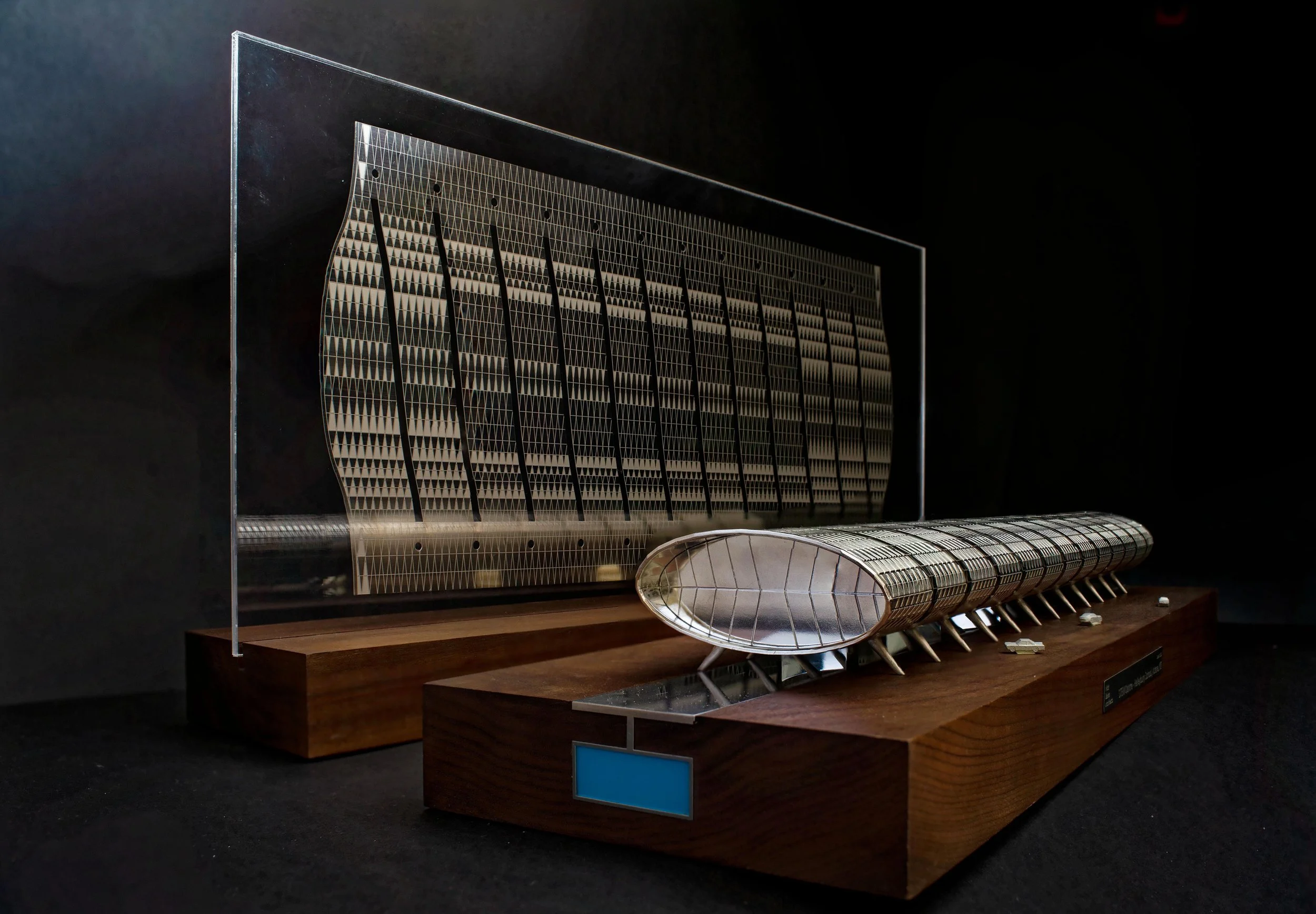 A detailed architectural model of a futuristic building with a curved grid structure, made of metal and glass, displayed on wooden stands in a dark exhibition setting.
