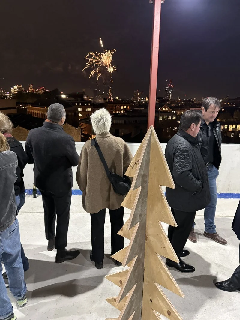 People watching fireworks in the night sky from a rooftop or terrace, with a wooden Christmas tree decoration in the foreground.