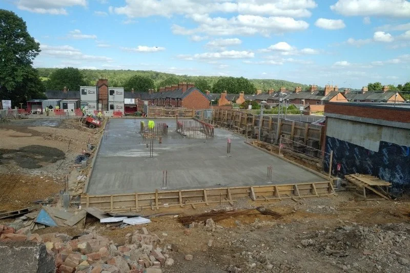 Construction site with freshly poured concrete foundation, surrounded by wooden formwork, in a residential neighborhood with houses and a partly cloudy sky in the background.